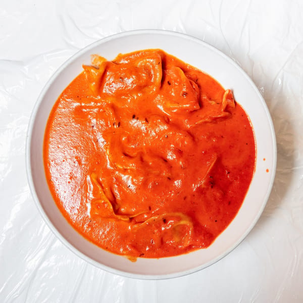 A white bowl filled with orange-red ravioli in a creamy sauce, on a white crinkled surface.